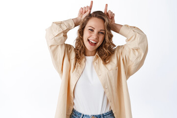 Naklejka premium Portrait of cheerful blond girl stay positive, showing stubborn horns gesture on head, smiling and looking happy, standing over white background