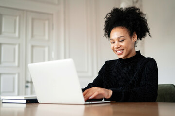 A satisfied female designer smiles at an Afro-ethnic appearance. A freelancer works remotely on a laptop in a browser on a new project for work. Curly black hair.