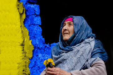 Grandmother with spring flowers on the background of the ruins. The old woman on the background of the slums. Portrait photo of a grandmother with flowers. Poverty and poverty. Helping the elderly