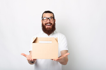 An excited young man with a delivery box is smiling amazingly at the camera near white wall