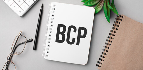 bcp is written in a white notebook with calculator, craft colored notepad, plant, black marker and glasses.