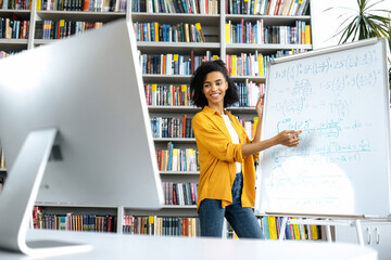 Stylish African American female coach, stands at whiteboard, in casual clothes, uses computer, leads an online lesson, communicates with students via video conference, looks at screen,distant learning