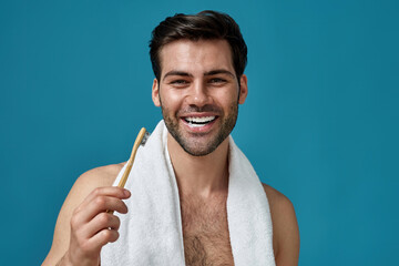 Portrait of happy bearded man with towel around neck smiling at camera with toothpaste in his mouth, holding wooden toothbrush isolated over blue background