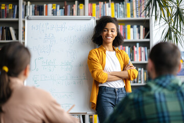 A young African American pleasant female teacher stands at the whiteboard in a university library...
