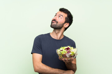 Young handsome man with salad over isolated green wall looking up while smiling