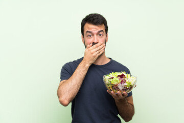 Young handsome man with salad over isolated green wall covering mouth with hands