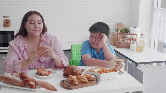 Asian family relationship of son upset with mother fat eating bread in the kitchen at home, mom hungry and eating bun with unhealthy food, two people, woman overweight, health care concept.