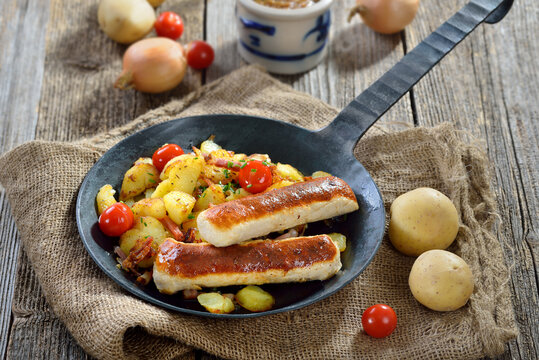 Gebratene bayerische Wollwürste mit Bratkartoffeln in der Eisenpfanne serviert – Roasted Bavarian sausages on fried potatoes with bacon, served in an iron frying pan 