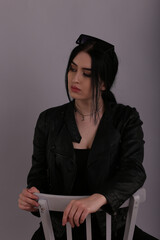 Young brunette woman in leather jacket sitting on a chair on grey background