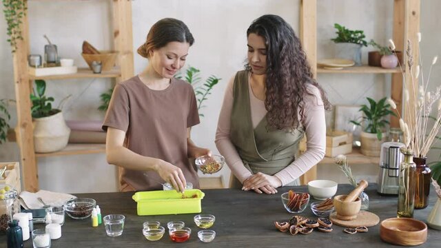 Medium PAN shot of two smiling women decorating and customizing handmade natural organic soap in silicon mold with dried floral petals