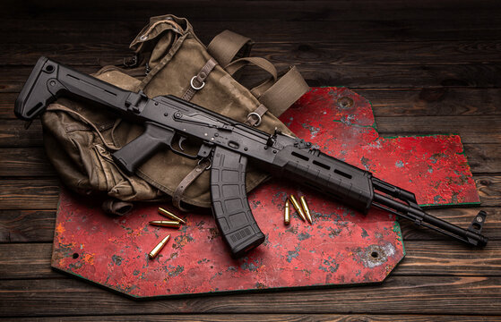 A classic Soviet military rifle AK in a modern body kit. The carbine and cartridges to it are on near the military backpack and the target for shooting. Weapons on a wooden table.