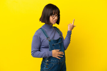 Young pregnant woman over isolated yellow background making the gesture of madness putting finger...
