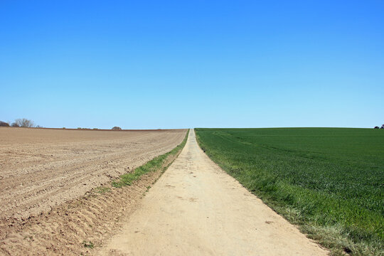 Three parts divided photo with blue clear sky, brown dry farmland and green growing grassland splitted by a straight way forward and horizon.