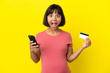 Young mixed race pregnant woman isolated on yellow background buying with the mobile and holding a credit card with surprised expression