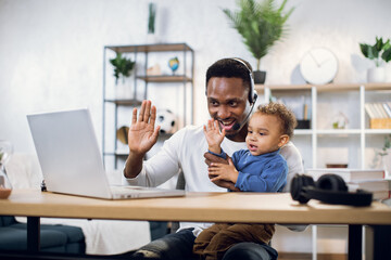 Afro american businessman with his little son on hands leading working meeting through video chat from home. Young father using modern laptop for work. - Powered by Adobe