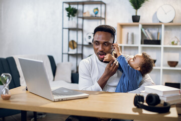 African man in headset having video chat on laptop while sitting at table. Young father trying to...