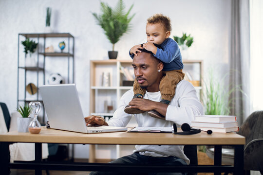 Afro american man carrying little son on neck, sitting at table and typing on modern laptop. Concept of freelance, multitasking and fatherhood.