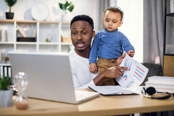 Afro american businessman having online meeting on modern laptop while carrying his baby son on...