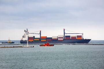 container cargo ship arrives at the port with the export goods delivered by nautical, escorting vessel by a tug boat at sea in cloudy weather.
