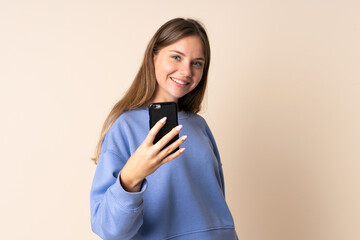 Young Lithuanian woman using mobile phone isolated on beige background with happy expression