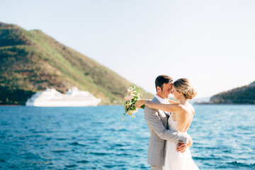 The bride and groom are embracing on the pier in the Bay of Kotor, behind them are green mountains and a white tourist ship 