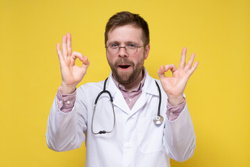 Contented, joyful doctor shows ok with both hands and smiling with a funny facial expression looks into the camera. Yellow background.