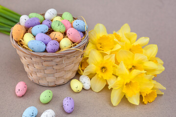 Basket with multi-colored Easter eggs stands on a beige background near a bouquet of daffodils. The concept of the holiday of Easter, Christ is risen. Place for text. close-up. Banner.