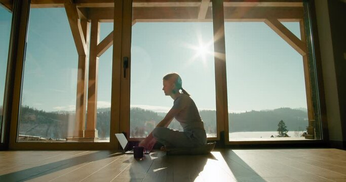 Young Lady Uses Laptop at Home. Woman Surfs Internet Sitting on Floor by Window with Mountains View and Drinks Tea in Sunny Day. Self-Isolation or Freelance Job Concept. Cinematic 4K Wide Zoom In Shot