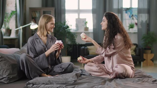 Medium PAN shot with slowmo of two young pretty women in silk pajamas sets sitting on bed in nice living room enjoying beauty routine together, smelling skincare products and chatting