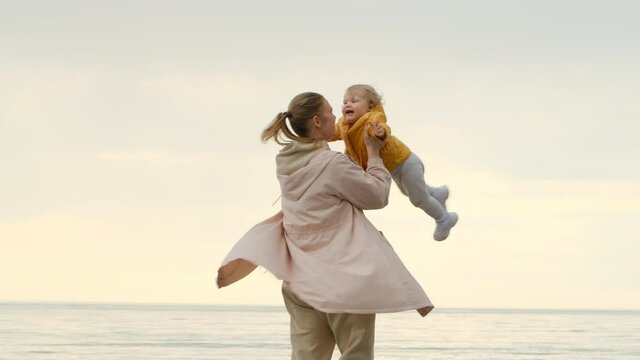 Happy mom and daughter on background of sea or ocean at sunset. mother turns or spin her daughter around her. Mom is wearing pink coat, and girl is wearing sweatpants and yellow raincoat.