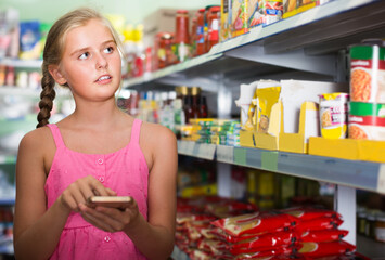 Portrait of blonde girl teenager touch phone screen and selects products in the supermarket