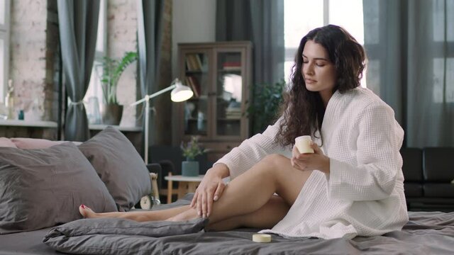 Medium PAN shot of young attractive brunette woman relaxing after bath in morning sitting on bed and applying moisturizing body lotion on her slim legs