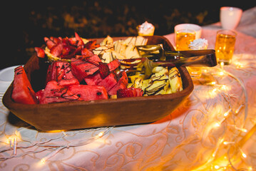 Wood fountain with fruit salad (watermelon, pineapple and strawberry) and chocolate sauce over a table with decoration and lights in a wedding