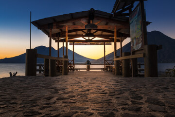 a lonely man in the middle of a sunrise waiting in a wooden shed on the shore of a lake with a background of a beautiful cloudscape and mountains