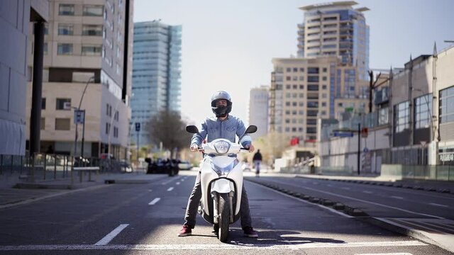 Wide shot of a young motorcyclist stopped at a traffic light in Barcelona. The man riding his scooter through the city on a large avenue lined with skyscrapers is waiting at the traffic light.