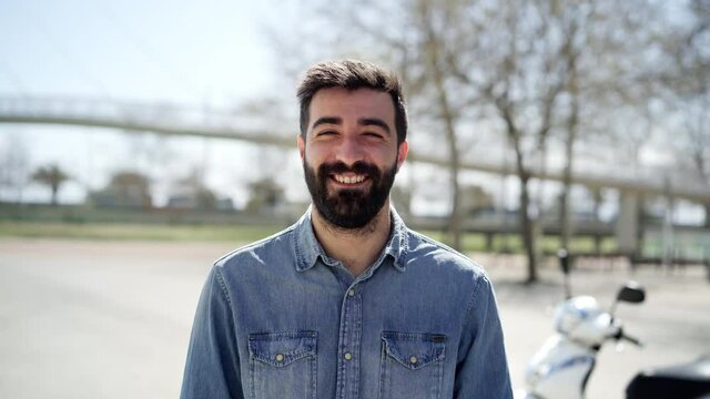 Portrait of a young Spanish man looking at the camera. The man with cheerful smile is in front of his motorcycle, which is parked in the city.