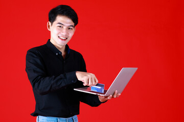 Asian young man smart and handsome do shopping online holding credit card and laptop on hand. He look happy with smile on face at studio with isolated red background. In concept shopping online