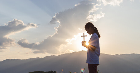Silhouette of human praying and holding christian cross for worshipping God at sunset background....