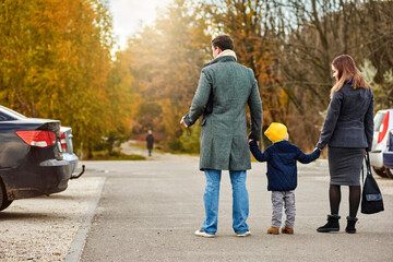 Family of three people walking to their parked car after stroll in autumn park