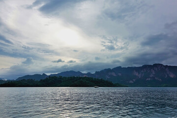 There are ripples on the surface of the lake. A boat is floating on the water. On the shore of a mountain with steep slopes. There are picturesque clouds in the sky. Cheow Lan Lake. Thailand