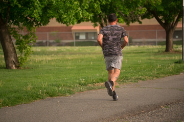 man wearing camouflage shirt and shorts running walking through park trail with green grass from behind wapato washington yakima county