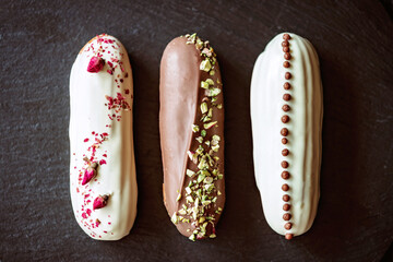 French dessert eclairs or profiteroles with chocolate icing and different toppings on a black stone plate. Pastry custard cakes with cream. Soft selective focus.