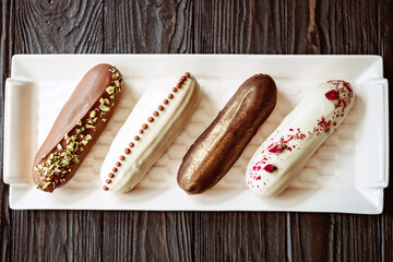 French dessert eclairs or profiteroles with chocolate glaze, with different toppings on a white plate, wooden background. Pastry custard cakes with cream. Soft selective focus.