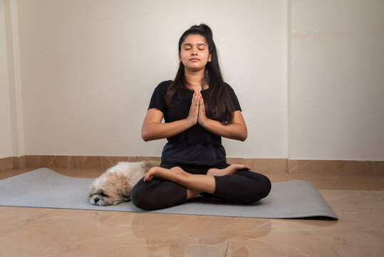 Young girl with closed eyes meditating on yoga mat on living room with her pet - concept of self-care, wellbeing and healthy lifestyle of millennials at home during coronavirus covid-19 lockdown.