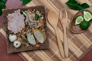 Dried tom yum noodles on dark plate wooden. Wooden spoon and fork. Garnish with the fruit and leaves of bergamot. Placed on a mat floor and an orange tiled backdrop.