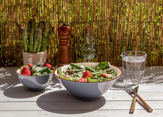 Fresh strawberry, green asparagus, feta cheese, baby spinach, and arugula salad served in a bowl with sparkling water. natural summer light and trendy strong reflection. Seasonal lunch on the balcony.