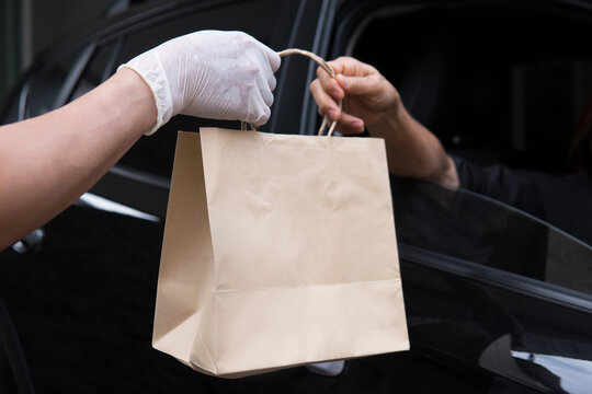 Closeup Food Delivery Man Hand Wear Glove Passing Brown Paper Bag To Woman Customer Driving Thru Pickup Out Of Black Car Window Meeting Social Distancing Requirements And Supporting Small Businesses