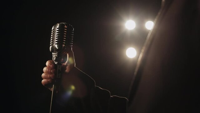 The singer is taking a retro microphone with his hand. The spotlight is shining on him. He's wearing a suit. Close-up shooting. 4K