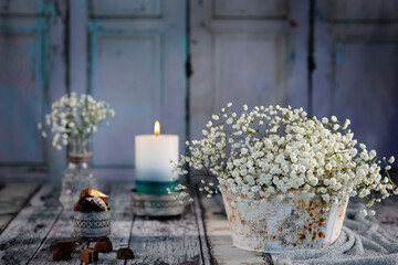 Blumenstrauß aus der weißen Schleierkraut steht in der antik Metallvase, im Hintergrund Glasvase auf dem alten Holz dekoriert mit dem Graustoff. Hintergrund ist hellblau, alte Holztür. Kerze brennt.