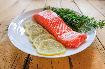 A piece of fresh trout fillet with lemon and dill greens on a white plate on a wooden table waiting for cooking. Restaurant menu. Dietary seafood. Keto and paleo diets. The concept of healthy eating.